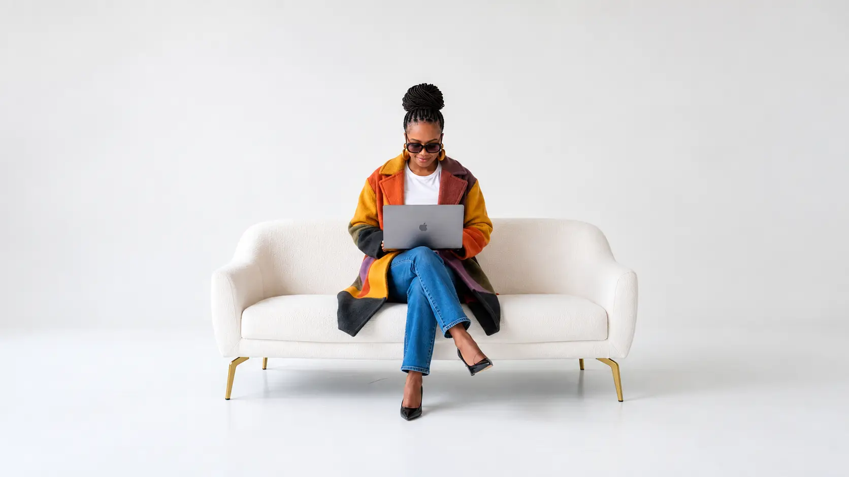 Raiona Denise sitting on a white couch in a colour-blocked coat, working on a laptop.